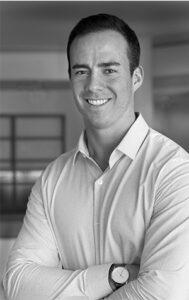A man with short dark hair smiles confidently at the camera, wearing a light-colored button-up shirt and a wristwatch, standing with arms crossed in a modern indoor setting.
