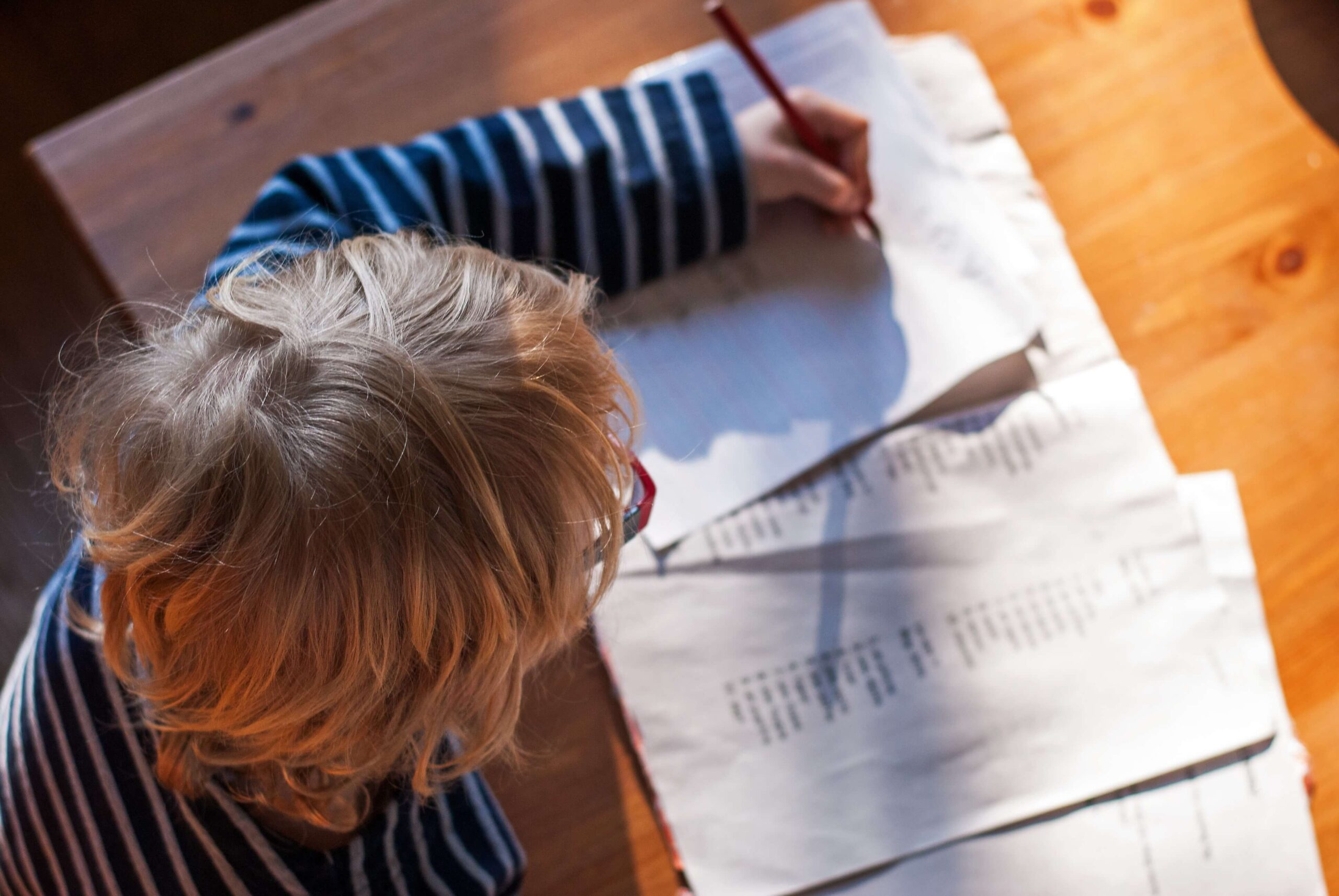 Brain Connections (1) A child with blonde hair wearing a striped shirt sits at a wooden table, writing with a pencil on a sheet of paper, with several papers spread out in front of them.