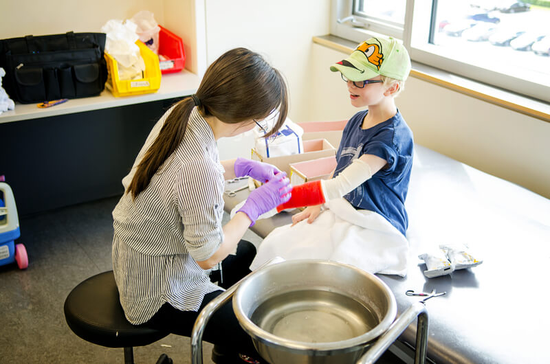 CIMT (1) A healthcare worker with purple gloves wraps a red cast on a young boy’s arm as he sits on an exam table, wearing glasses and a green cartoon hat. Medical supplies and a large metal bowl are nearby.