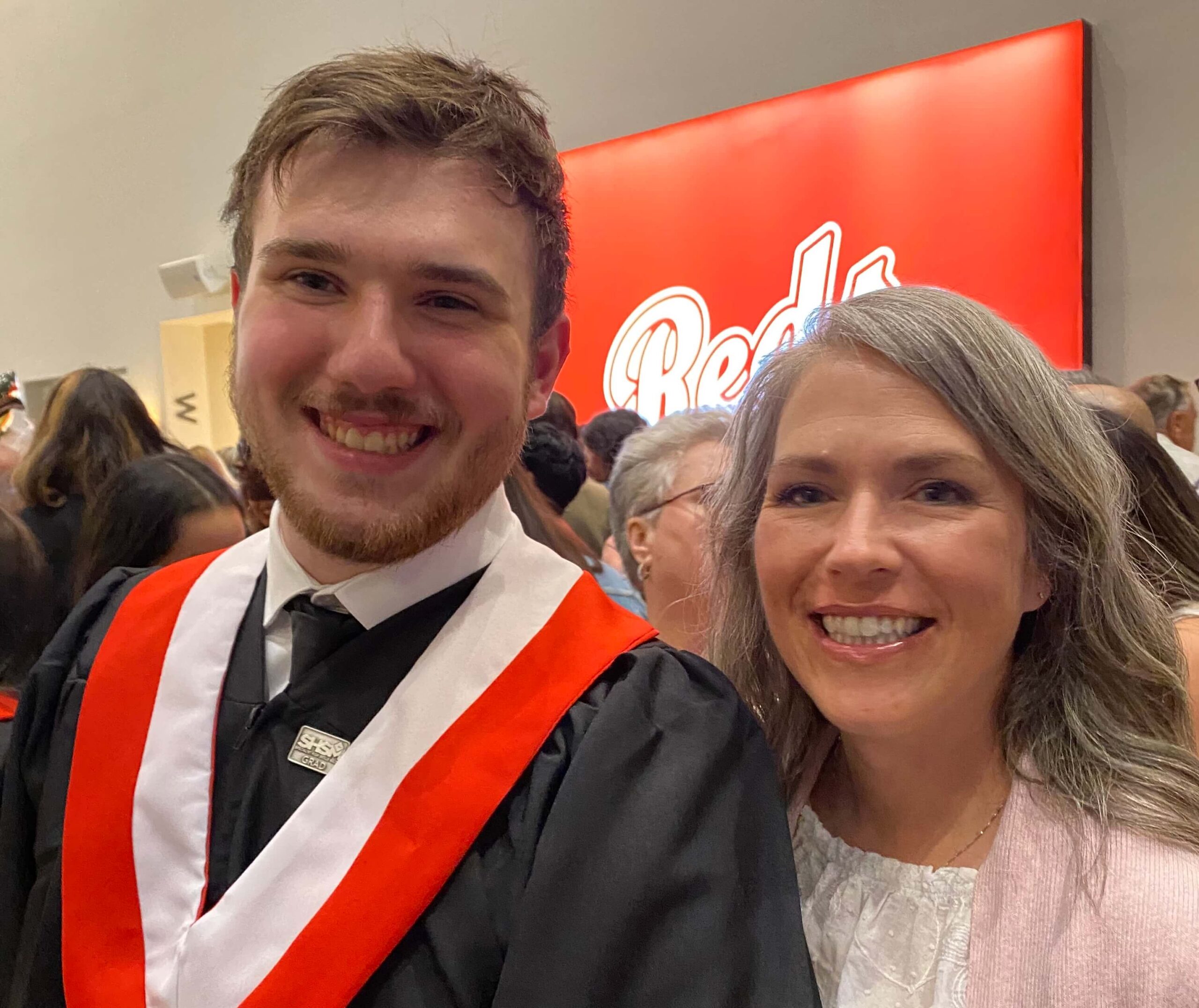 A young man in a graduation gown with red and white trim smiles next to a woman with gray hair. They are indoors with a red Reds sign and a crowd of people in the background.