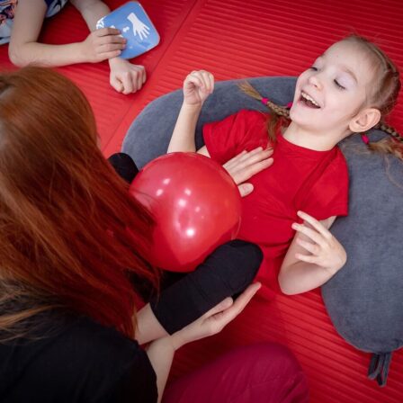 A young girl with braided hair lies on a gray pillow, smiling and looking up while holding a red balloon. An adult sits beside her, engaging with her. Both are on a red padded mat.