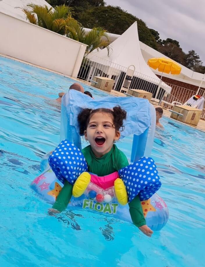 A young child in a green swimsuit smiles joyfully while floating in a pool using a blue polka-dot floatation device, with palm trees, a white fence, and people in the background.