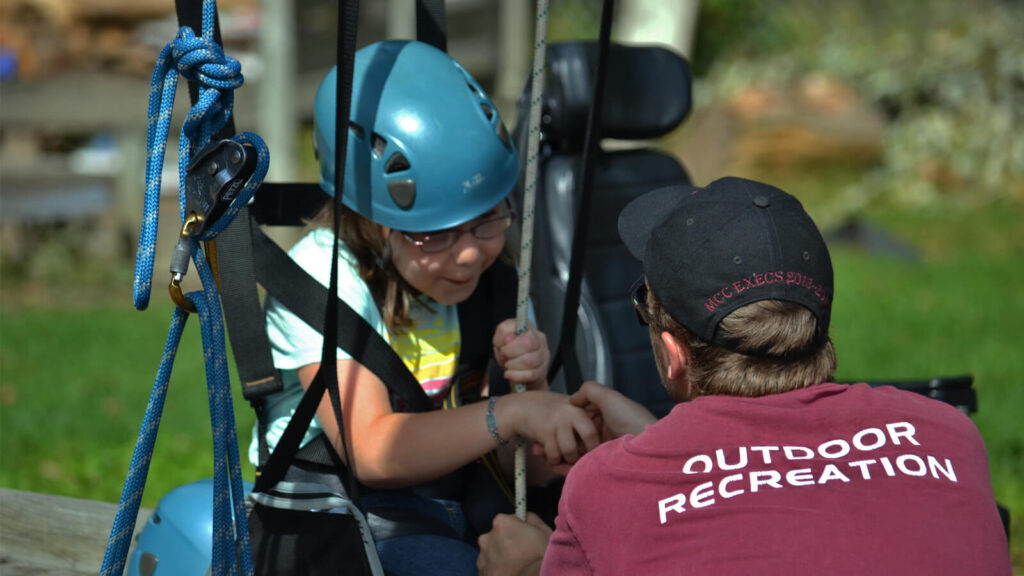 A young girl wearing a helmet sits in a harness swing, holding a rope, while an instructor in a maroon “Outdoor Recreation” shirt kneels beside her, offering guidance and support outdoors.
